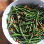 Top down image of a serving bowl filled with green beans almondine, bright green beans garnished with golden fried almonds, shallots, garlic and lemon zest.