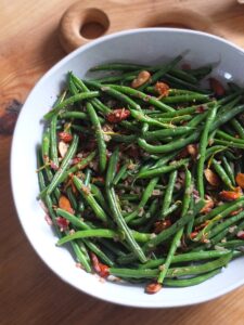 Top down image of a serving bowl filled with green beans almondine, bright green beans garnished with golden fried almonds, shallots, garlic and lemon zest.