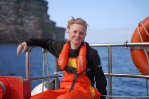 Image of Elizabeth Atia on the fishing boat Boy Frazer II with the cliffs of Noss in the background. Image was taken after a day spent fishing mackerel and filming for the Channel 5 tv series Shetland: Scotland's Wondrous Isles. Elizabeth's face and clothes are splattered with mackerel scales, she's tired, but she's still smiling.